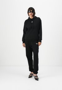 Black hoodie with a front logo, paired with black joggers. Both items feature a smooth texture. Worn with pointed black heels.