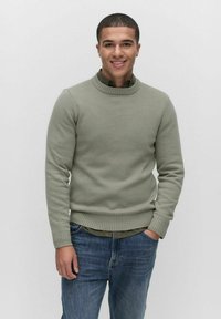 Ljusgrön stickad tröja med hög ribbad krage och ribbade ärmar, i kombination med en mörkgrön skjorta med krage under och blå jeans.