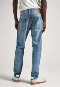 Pequeños jeans de mezclilla azul claro con corte recto, que presentan dos bolsillos traseros y un parche de cuero marrón en la cinturilla. Zapatillas deportivas blancas informales visibles.