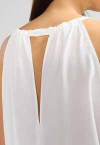 Blusa blanca con un agujero en la parte posterior y cierre de botón, fabricada en un tejido suave y con un diseño de corte holgado. Textura suave y estilo minimalista.