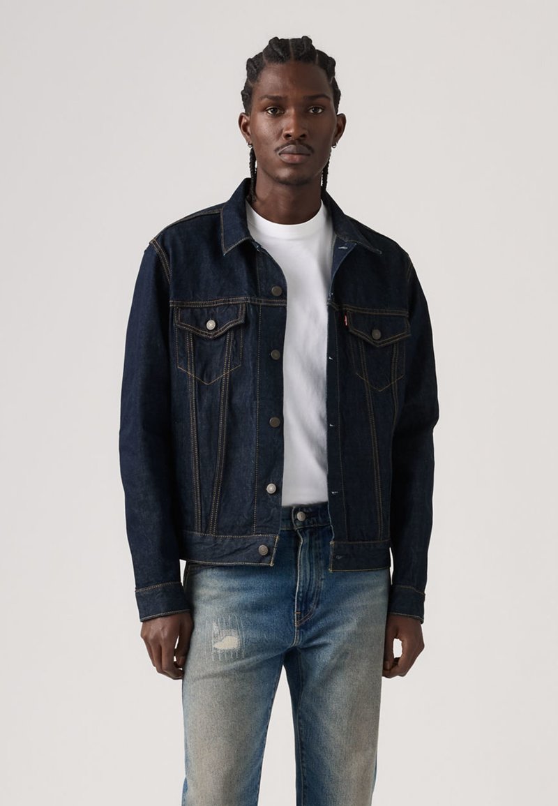 Dark blue denim jacket with button closures, two chest pockets, and contrasting stitching. Worn over a white T-shirt and light blue jeans.