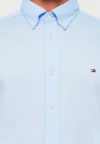  Lichtblauw overhemdd met een kraag, voorzien van witte knopen en een klein geborduurd logo in rood, wit en blauw op de borst.