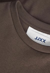 Smeđi pamučni dukserica s rebrastim okruglim izrezom. Unutarnja etiketa sadrži tekst "JJXX" u plavoj boji. Glatka tekstura, minimalistički dizajn.