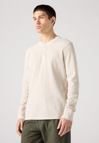 Langermet, ribbestrikket, beige henley-skjorte med tre knapper ved kragen. Myk tekstur, figurtilpasset design og standardmansjetter. Kombinert med grønne bukser.