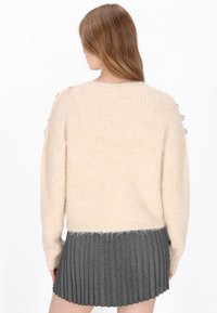 Maglione beige a maglia con una texture a coste, caratterizzato da un orlo inferiore peloso, abbinato a una gonna plissettata scura. Nessun motivo o accento visibile.