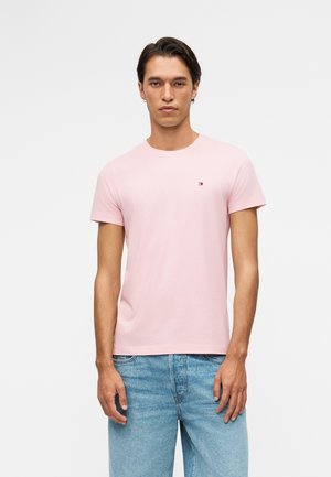 Mężczyzna w jasno różowej koszulce z krótkim rękawem i jasnoniebieskich dżinsach, stojący na jednolitym białym tle.