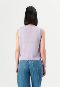 Person med skulderlangt mørkt hår iført enermet lavender strikket topp og blå denimjeans, stående med ryggen til på en hvit bakgrunn.