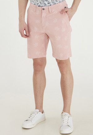 Helle rosa Shorts aus Baumwolle mit einem Motiv von Palmen, seitlichen Taschen und einem Knopfverschluss. Getragen mit weißen Turnschuhen.