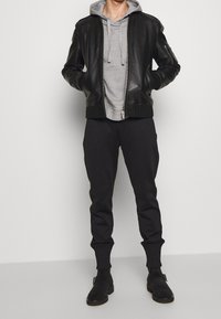 Chaqueta bomber de cuero negro sobre un hoodie gris, pantalones de chándal negros ajustados y zapatos negros sin cordones. Los materiales son suaves y de textura lisa.