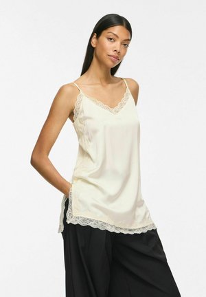 Femme aux longs cheveux foncés portant un caraco crème orné de dentelle et un pantalon noir ample, debout avec une main dans la poche.