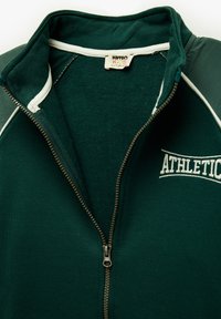 Grüne Zip-Up-Sweatjacke mit einem Fleece-Innenfutter, weißen Akzenten entlang des Reißverschlusses und der Aufschrift "ATHLETIC" auf der Vorderseite.