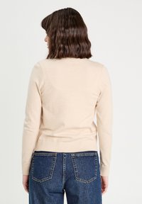 Pull en tricot beige à manches longues, col rond, ourlet côtelé, porté sur un jean en denim bleu, vu de dos.
