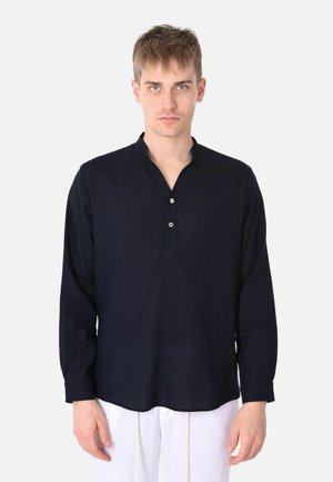 Giovane uomo con capelli corti castano chiaro che indossa una camicia blu scuro a maniche lunghe con bottoni e pantaloni bianchi su uno sfondo uniforme.