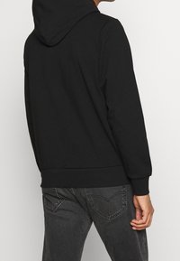 Felpa nera con cappuccio regolabile, polsini a coste, tessuto morbido e forma dritta, abbinata a jeans scuri con tasche posteriori cucite.