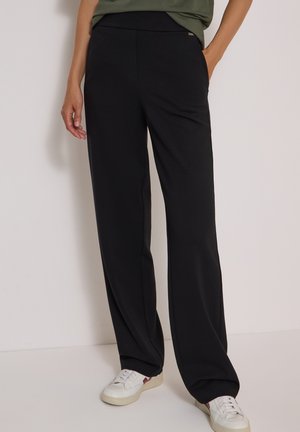 Femme portant un pantalon noir ample et des baskets blanches, debout avec une main dans la poche devant un mur uni.