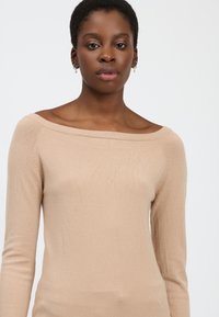 Maglione beige off-shoulder con maniche lunghe, texture morbida e design aderente. Dettagli sartoriali sottili lungo le spalle e le maniche.