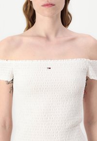 Valge õlgadelt avatud pluus, valmistatud tekstureeritud kangast koos kokku tõmmatud detailidega. Ees on väike tikitud logo.