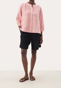 Blusa a rayas rojas y blancas con botones y escote redondo, combinada con shorts negros. Lleva sandalias de dedo marrones y tiene un corte relajado.