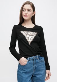 Črna majica z dolgimi rokavi z izrezanim trokotnim logotipom "GUESS" iz srebrnih in kremnih sequinsov, kombinirana s svetlo modrimi visokim pasom jeans hlačami.