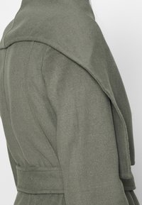 Cappotto verde oliva con un ampio colletto a scialle, texture morbida, vita cintata e silhouette sagomata, che enfatizza i contorni del design.