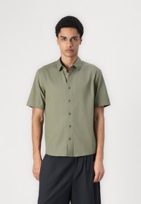 Chemise à manches courtes boutonnée en tissu vert olive. Présente un col droit, sept boutons et une coupe décontractée. Design minimaliste.