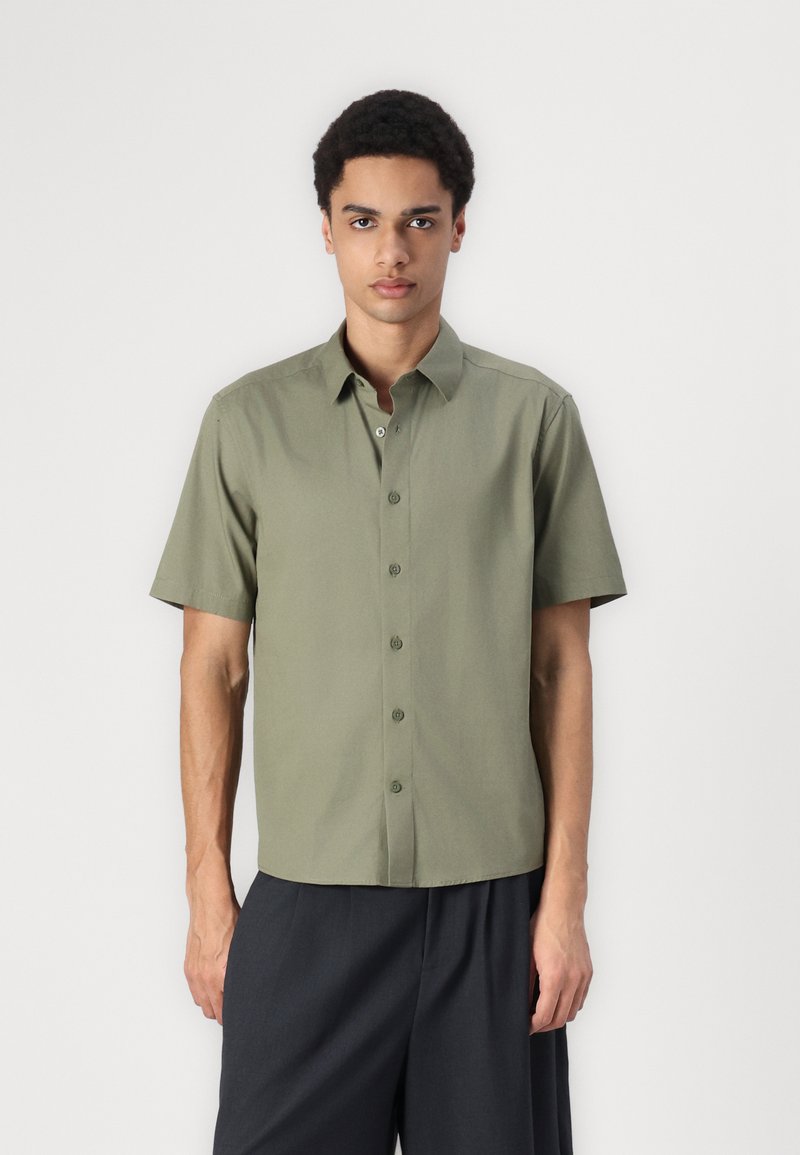 Chemise à manches courtes boutonnée en tissu vert olive. Présente un col droit, sept boutons et une coupe décontractée. Design minimaliste.