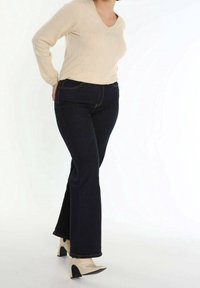 Beżowy sweter dzianinowy z dekoltem w serek, w połączeniu z ciemnoniebieskimi spodniami dzwonami. Modelka nosi kremowe lakierowane buty na obcasie z czarnymi akcentami.