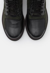 Botas de tobillo de cuero negro y gamuza verde con punta redonda, diseño de cordones y detalles de costura roja en contraste.