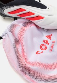 Bijele nogometne kopačke s crnim i crvenim naglascima pored torbe na vezanje koja ima gradijentni uzorak s ružičastim i crnim točkicama te natpis "COPA PURE III".