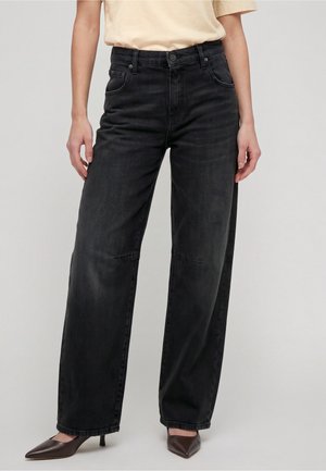 Kvinde iført sorte jeans med vide ben, beige kortærmet top og brune spidse hæle, står foran en ensfarvet baggrund.