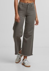 Pantalon à jambes larges en denim vert olive, avec un ourlet effiloché, un design classique à cinq poches et une fermeture avant par bouton. Associé à des baskets blanches.