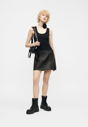 Jeune femme aux cheveux blonds bouclés portant une robe noire sans manches, des bottes noires, un collier ras du cou noir avec une fleur et tenant un sac à bandoulière noir.