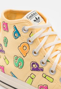 Höga sneakers i gul canvas med färgglada, djärva bokstäver tryckta på sidorna. Vita snören och metallhål för fastsättning.