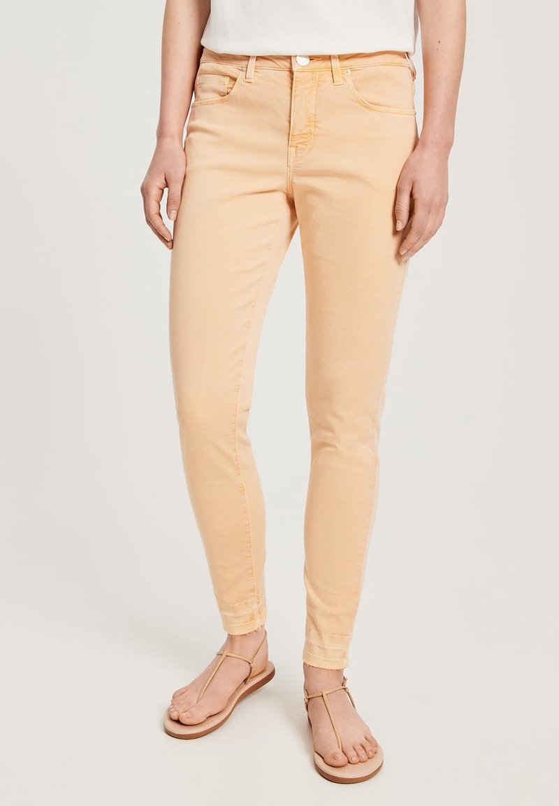 Jeans ajustados de color melocotón hechos de denim, con una cinturilla estándar, bolsillos frontales y detalles de costura sutiles. Llevados con sandalias planas.