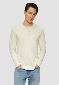 Cremfarbener Langarm-Henley-Pullover mit drei goldenen Knöpfen am Halsausschnitt, kombiniert mit hellblauen Jeans. Weicher Stoff und figurnaher Schnitt.