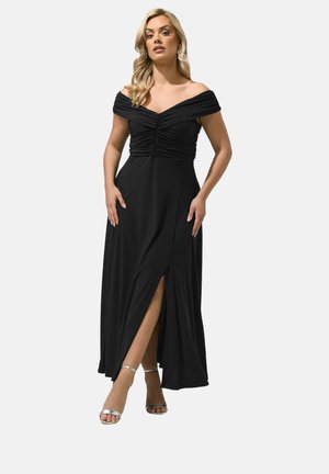 Vrouw draagt een zwarte off-shoulder jurk met geplooide stof en een split aan de voorkant, en zilveren open tenen hakken, staand met handen langs het lichaam tegen een effen achtergrond.