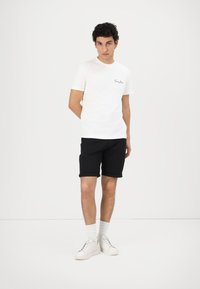T-shirt blanche à manches courtes avec un petit logo sur la poitrine, associé à un short noir à ourlet roulé et des baskets blanches, se tenant contre un fond uni.