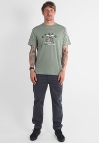 T-shirt en coton vert avec un motif à l'avant représentant un vélo et une planche de surf. Porté avec un pantalon gris foncé et des baskets noires.