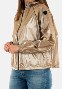 Giacca metallic beige con cappuccio, chiusura con zip, polsini elasticizzati e una toppa rotonda con logo sulla maniche sinistra. Texture liscia e lucida.