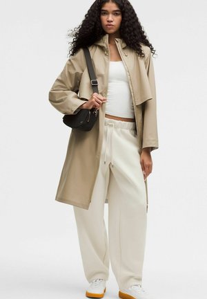 Młoda kobieta o kręconych włosach, ubrana w beżowy trencz, biały top na ramiączkach, luźne białe spodnie, białe trampki z pomarańczowymi podeszwami oraz czarną torebkę na ramię.