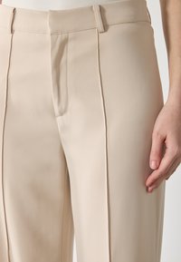 Beige op maat gemaakte broek met een slank ontwerp, soepele stof en voorplooien. Een hand rust lichtjes op het dijgebied.