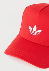 Κόκκινο καπέλο από λείο ύφασμα με λογότυπο τριών λευκών ρίγων και επίπεδη γείσο. Οι ραφές είναι ορατές κατά μήκος των άκρων.