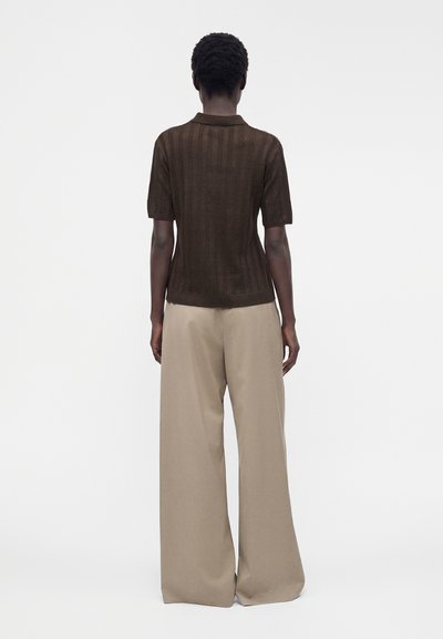 Personne debout de dos, portant un t-shirt côtelé marron foncé à manches courtes et un pantalon beige à jambes larges sur un fond uni.