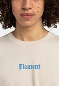 Beige cotton t-shirt featuring bold blue "Element" text design, crew neckline, and smooth fabric texture.