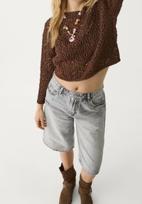 Top corto de punto marrón con tejido abierto, combinado con pantalones cortos Bermuda de denim gris claro. Botas de tobillo marrones acentúan el atuendo.