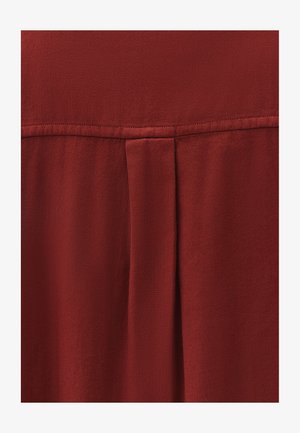 Tessuto rosso, testurizzato, con motivo a spina di pesce. Presenta un dettaglio a plissettatura verticale e una cucitura piatta all'ascella. Morbido e cade delicatamente.