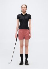Γυναίκα που στέκεται με μπαστούνι γκολφ, φοράει μαύρο πόλο μπλουζάκι με κοντά μανίκια, ροζ σορτς, μαύρες κάλτσες και μαύρα παπούτσια σε απλό φόντο.