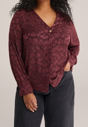 Kobieta ubrana w bordową, teksturowaną bluzkę z długim rękawem z pojedynczym ozdobnym guzikiem oraz czarne spodnie, z ręką w kieszeni.