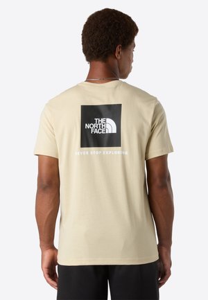 Mand iført en beige t-shirt med et sort firkantet logo og teksten "The North Face" samt sloganet "Never Stop Exploring" på ryggen.