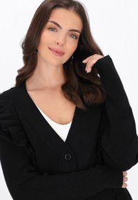 Fekete bordázott kardigán fodros részletekkel az ujjak mentén, mély V-nyakkal és egy fekete gombos záródással. Puha tapintás és testhez simuló forma.